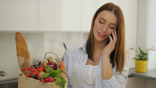 Jonge vrouw praten over haar mobiele telefoon na het winkelen in de keuken — Stockvideo