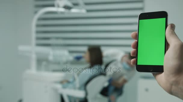 Smartphone mit grünem Bildschirm, vor dem Hintergrund des arbeitenden Zahnarztes und Patienten. — Stockvideo
