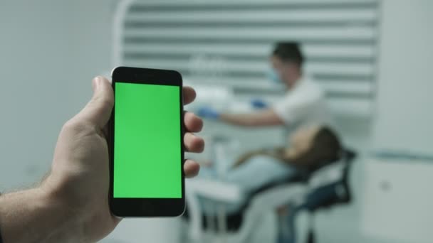 Smartphone mit grünem Bildschirm, vor dem Hintergrund des arbeitenden Zahnarztes und Patienten. — Stockvideo