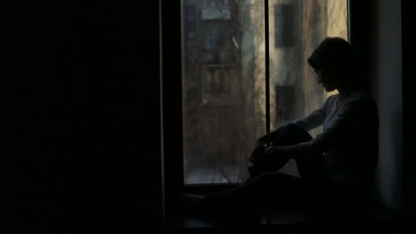 Pervazın üzerinde oturan ve pencereden dışarı bakarak üzgün kadın silüeti — Stok video