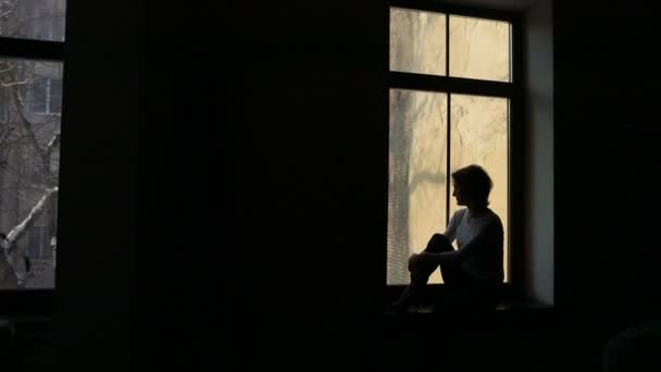 Sylwetka smutna kobieta siedzi na parapet i patrząc przez okno — Wideo stockowe