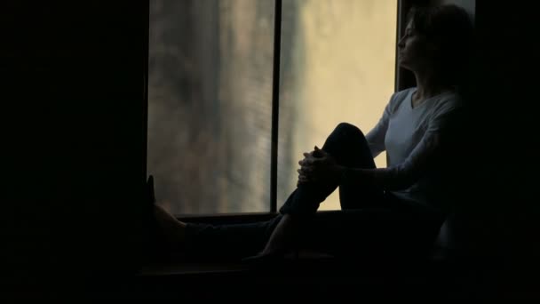 Силуэт женщины, сидящей на подоконнике и смотрящей в окно — стоковое видео