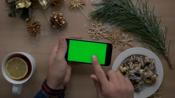 Noel dekorasyonu ile ışık ahşap zemin üzerine yeşil ekranlı akıllı telefon kullanarak ele mans. Üstten Görünüm. Chroma anahtar. Yatay konumu. — Stok video