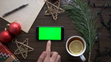Bir şey çay, yıldız, oyuncak ve garland kahverengi ahşap zemin üzerine yeşil ekranlı akıllı telefon arama el mans. Üstten Görünüm. Chroma anahtar. Yatay konumu.