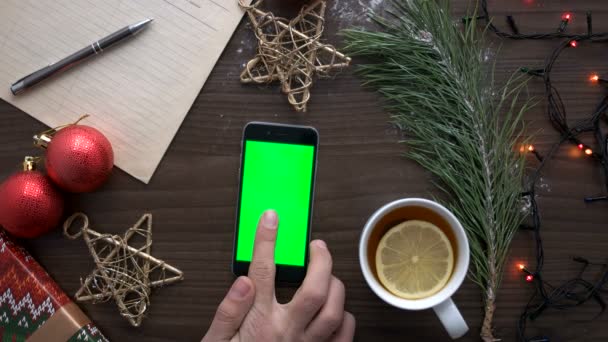 Чоловік шукає щось у смартфоні з зеленим екраном на коричневому дерев'яному фоні з чаєм, зірками, іграшками та гірляндою. Вид зверху. Клавіша Chroma . — стокове відео