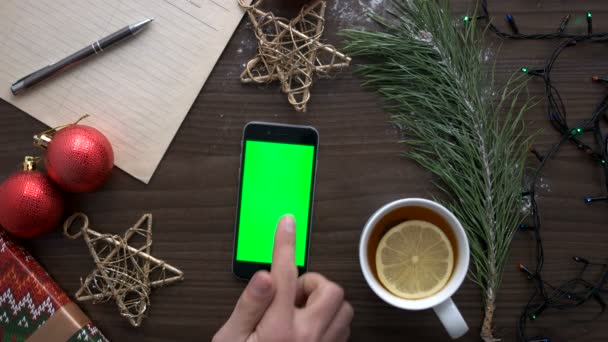 Mans hand using smart phone with green screen on brown wood background with tea, stars, toys and garland. Вид сверху. Ключ хрома . — стоковое видео
