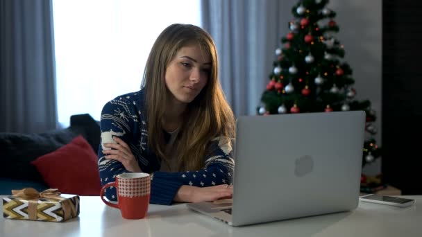 Allvarlig ung kvinna placering på bordet och använder laptop på julgran bakgrunden. — Stockvideo