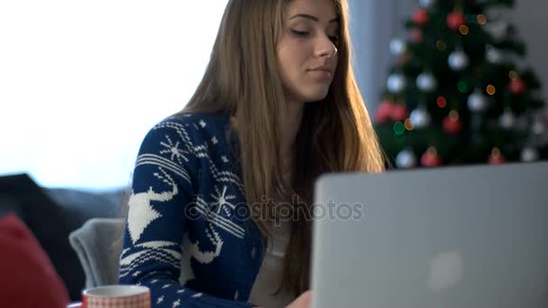 Mosolygó fiatal nő, írja be a laptop, és beszél a nappaliban a karácsonyfa háttér szúró telefon. Közelről. — Stock videók