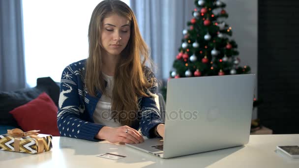 Serieuze jonge vrouw aanbrengen op de tafel en de kopie de cijfers van kaarten naar de laptop op de achtergrond van de kerstboom. — Stockvideo