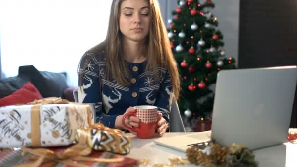 Kobieta pije kawę przy stole z świątecznych dekoracji i laptop na tle choinki. — Wideo stockowe