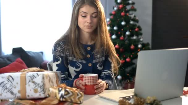 Vrouw koffie drinken aan de tafel met Kerstdecoratie en laptop op de achtergrond van de kerstboom in de woonkamer. — Stockvideo