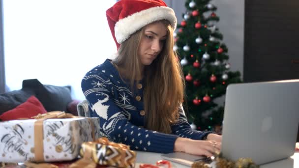 Mujer joven seria en el sombrero de Santa Claus sentado en la mesa y el uso de la computadora portátil en el fondo del árbol de Navidad . — Vídeos de Stock