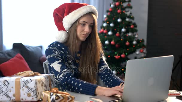 Souriant jeune femme dans Santa Claus chating avec un ami sur ordinateur portable et boire du café sur le fond de l'arbre de Noël dans le salon . — Video