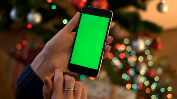 Vrouw handen verticale smartphone met groen scherm op de achtergrond met bokeh van garland. Close-up. Chromakey. — Stockvideo