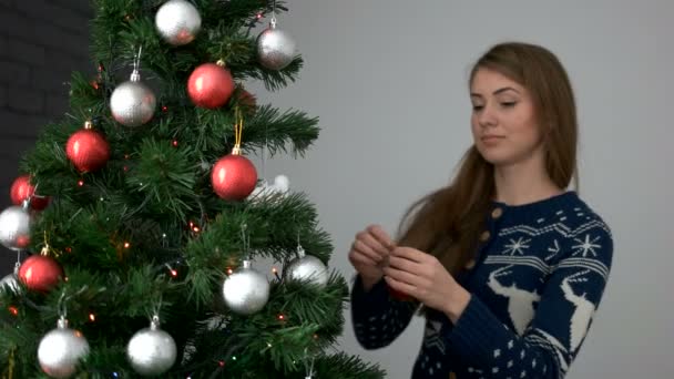 Όμορφη νεαρή γυναίκα με τα πόδια από το δέντρο, διακοσμώντας το χριστουγεννιάτικο δέντρο με κόκκινο, ασημί παιχνίδια σε γκρίζο φόντο. — Αρχείο Βίντεο