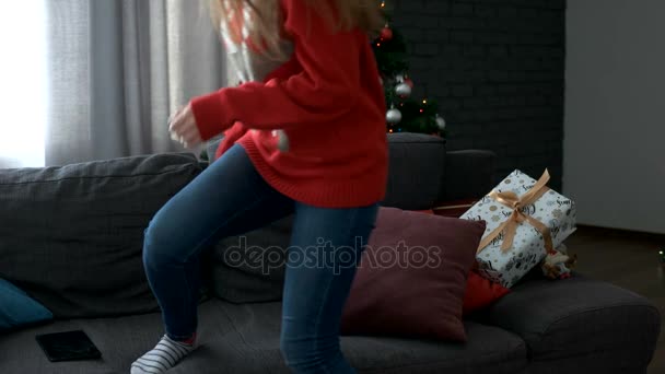 Jonge aantrekkelijke gelukkige vrouw komt naar de woonkamer, zittend op de Bank met gave en start met het gebruik van Tablet PC op kerstboom achtergrond. — Stockvideo