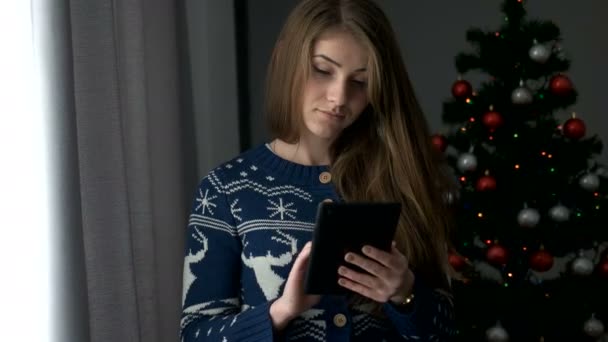 Porträt einer jungen attraktiven Frau, die auf einem Tablet auf Fensterhintergrund mit Weihnachtsbaum tippt. Nahaufnahme. — Stockvideo