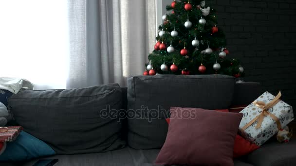 Jovem mulher atraente que vem para a sala de estar, sentada no sofá com presente e começar a escolher o presente em tablet no fundo da árvore de Natal . — Vídeo de Stock