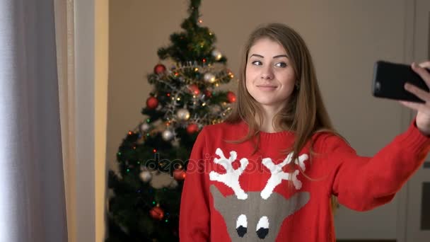 Jovem mulher em suéter com geer tomando selfie no telefone inteligente no fundo da árvore de natal. Jovem mulher atraente posando na câmera . — Vídeo de Stock