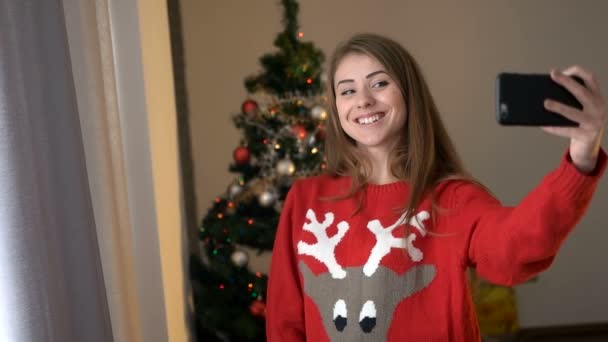 Mujer joven en suéter con geer tomando selfie en el teléfono inteligente en el fondo del árbol de Navidad, posando y sonriendo en la cámara . — Vídeos de Stock