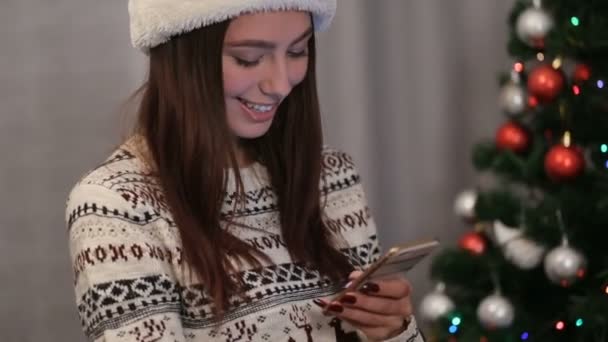 Mladá usmívající se žena v santa claus klobouk usmívá a posílání sms pozdravy pomocí chytrého telefonu na pozadí vánoční stromeček. — Stock video