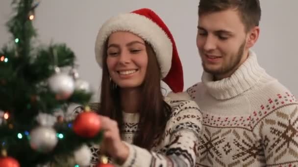 Feliz pareja joven decorando el árbol de Navidad en suéteres lindos. Pareja enamorada preparándose para la víspera de Navidad . — Vídeos de Stock