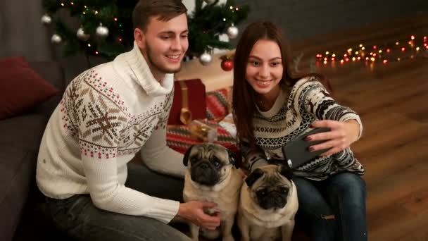 Glada unga par i kärlek leende och göra selfie med par mops hunden nära julgranen. Ovanifrån. — Stockvideo