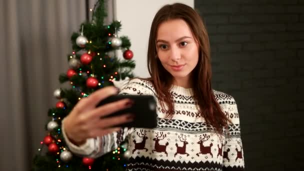 Attractive young woman taking selfie and posing on smart phone camera with christmas tree in sweater with deer and christmas prints. — Stock Video