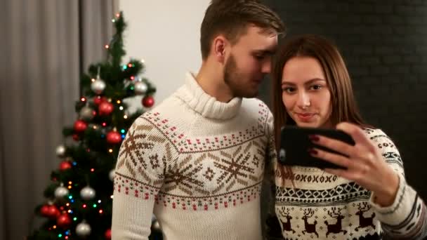 快乐的年轻男人和女人拥抱和以自拍照在圣诞节毛衣圣诞树背景上的智能手机. — 图库视频影像