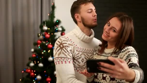 Feliz jovem casal emocional posando na câmera de telefone inteligente no fundo árvore de natal . — Vídeo de Stock