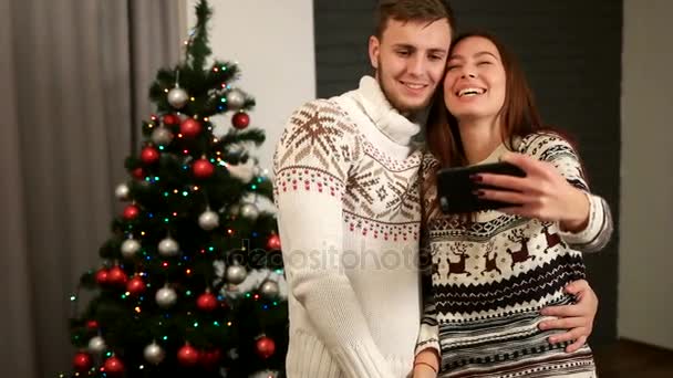 Feliz casal emocional no amor posando na câmera do telefone inteligente no fundo árvore de natal em camisolas de Natal . — Vídeo de Stock