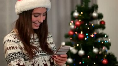 Akıllı telefon stending Noel ağacı yakınındaki kullanarak santa claus şapka genç çekici kadın.