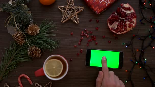 クリスマス平面図です。男の手が緑色の画面でスマート フォンを使用します。指タッチ スクリーンにページをスクロールします。クリスマス木製テーブルの背景についてクロマ キー。上からの眺め — ストック動画