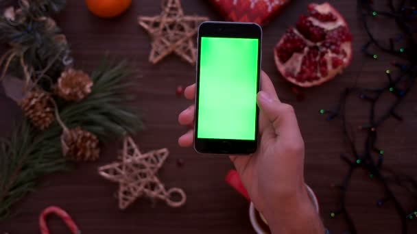 Vista superior de la mano del hombre sosteniendo el teléfono inteligente con pantalla verde. Dedo tocando y páginas de desplazamiento en la pantalla táctil en el fondo de la mesa de Navidad. Clave de croma — Vídeos de Stock