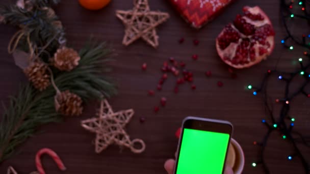 Vánoční pohled shora. Člověk ruku pomocí chytrého telefonu s zeleným plátnem. Posouvání stránky prst na dotykovém displeji. Vánoční podrobnosti o dřevěný stůl pozadí. Chromatický klíč. Pohled shora — Stock video