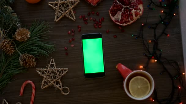 Рука людини зверху за допомогою смартфона з зеленим екраном. Палець прокручує сторінки на сенсорному екрані. Різдвяна прикраса на дерев'яному фоні столу. Клавіша Chroma — стокове відео