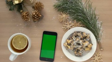 Noel üstten görünüm. Ahşap masa üstünde yeşil perde ile akıllı telefon. Noel dekorasyonu, bisküvi, çay. Chroma anahtar