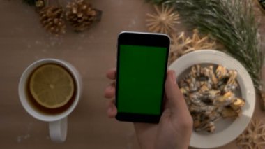 Noel üstten görünüm adam el yeşil ekranlı akıllı telefon tutarak. Dokunarak ve Noel masa artalanını dokunmatik ekran sayfalarında kaydırma parmak. Chroma anahtar
