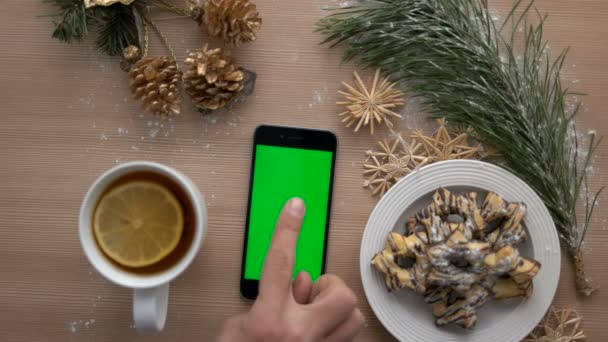 Bovenaanzicht man hand met behulp van slimme telefoon met groen scherm. Vinger te tikken en te scrollen van pagina's op touchscreen. Kerst decor op houten tafel achtergrond. Chromakey — Stockvideo