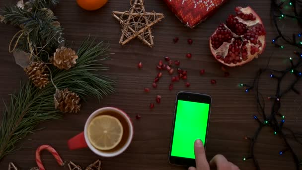 Manos de hombre de vista superior tocando en una pantalla táctil de teléfono inteligente con pantalla verde. Decoración navideña sobre el fondo de la mesa de madera. Clave de croma. Disparo desde arriba. — Vídeos de Stock