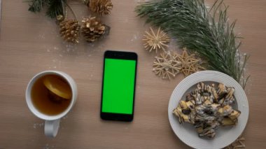 Noel üstten görünüm. Ahşap masa üstünde yeşil perde ile akıllı telefon. Noel dekorasyonu, bisküvi, çay. Chroma anahtar