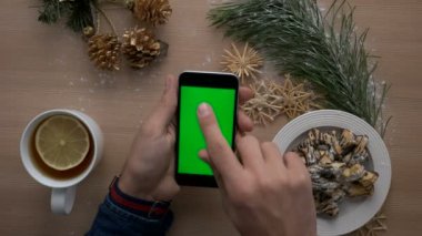 Noel dekorasyonu ile ahşap masa üzerinde yeşil ekranlı akıllı telefon kullanan adam. Rahat Noel. Üstten Görünüm. Chroma anahtar