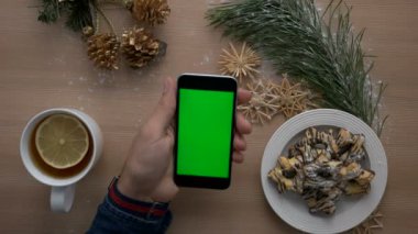 Noel dekorasyonu ile ahşap masa üzerinde yeşil ekranlı akıllı telefon kullanan adam. Rahat Noel ayrıntılar. Üstten Görünüm. Chroma anahtar