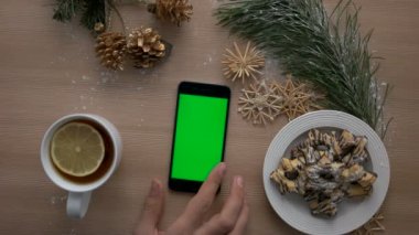 Noel dekorasyonu ile ahşap masa üzerinde yeşil ekranlı akıllı telefon kullanan adam. Adam sayfaları çevirme. Rahat Noel. Üstten Görünüm. Chroma anahtar