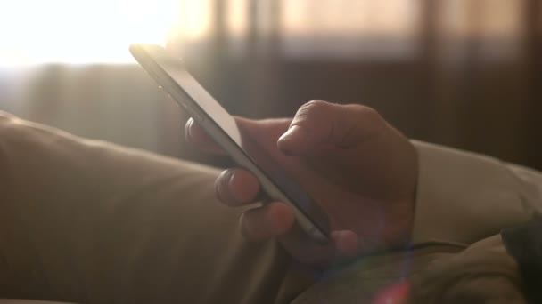 Feche as mãos masculinas segurando o telefone inteligente. Homem usando telefone celular enquanto sentado n o sofá em casa. Tocando na tela sensível ao toque — Vídeo de Stock