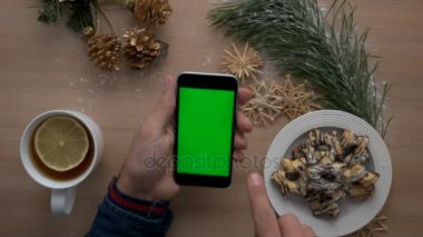 Noel dekorasyonu ile ahşap masa üzerinde yeşil ekranlı akıllı telefon kullanan adam. Rahat Noel ayrıntılar. Üstten Görünüm. Chroma anahtar