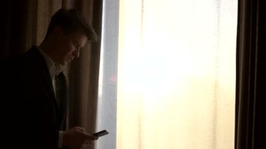 Genç işadamı evde pencere yanında bir smartphone kullanarak. Pencere duran ve cep telefonuyla genç adam silüeti.