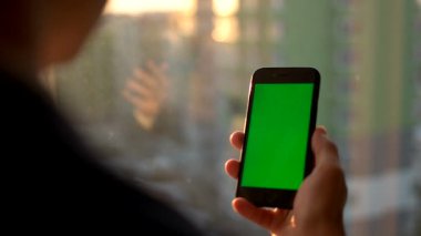 Erkek el yeşil ekranlı akıllı telefon tutarak. Pencere duran cep telefonu kullanan adam. Arkadan Görünüm vurdu. Chroma anahtar