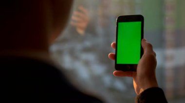 Erkek el yeşil ekranlı akıllı telefon tutarak. Pencere duran cep telefonu kullanan adam. Arkadan Görünüm vurdu. Chroma anahtar