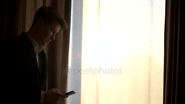 Joven hombre de negocios usando un teléfono inteligente al lado de la ventana en casa. Silueta de hombre joven de pie cerca de la ventana y el uso de teléfono móvil . — Vídeo de stock